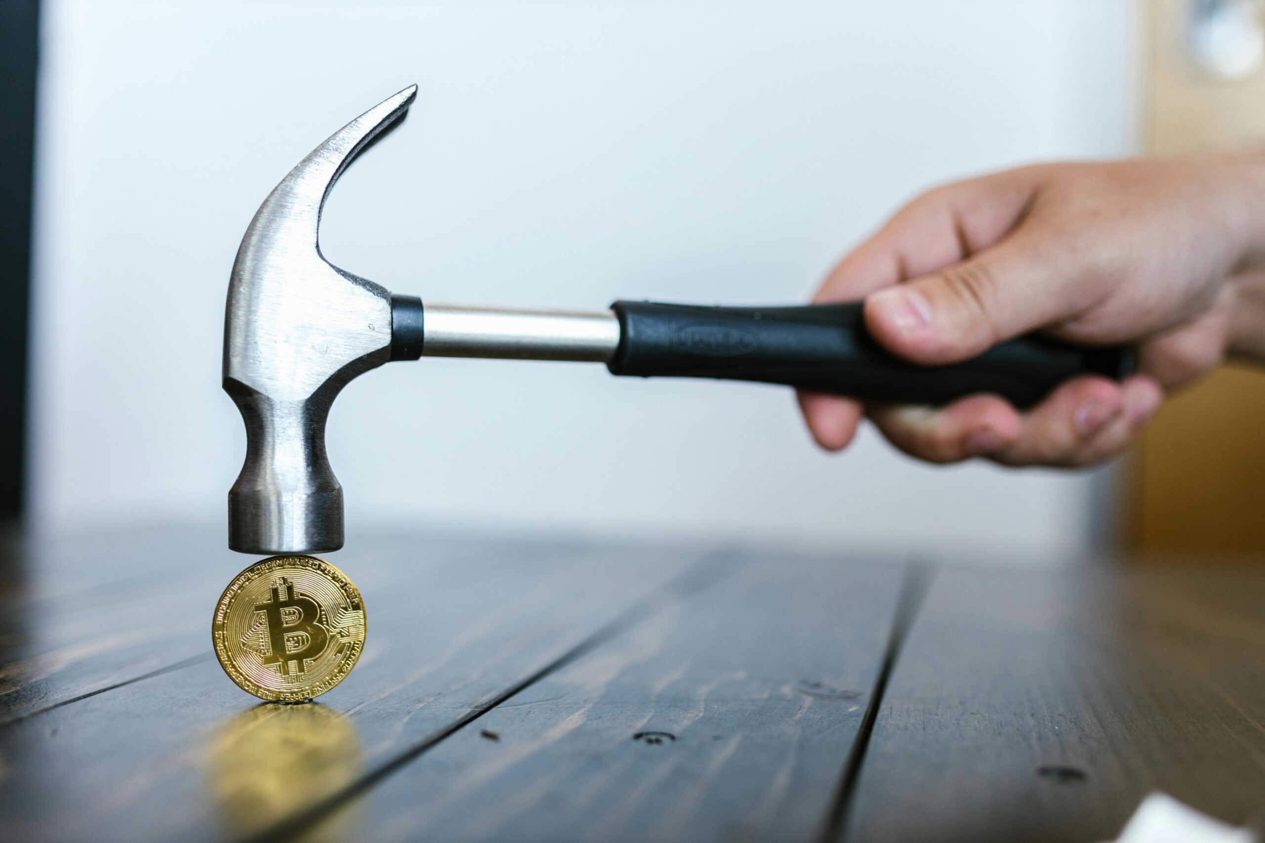 A hand holding a hammer above a Bitcoin coin on a wooden surface, symbolizing cryptocurrency volatility.